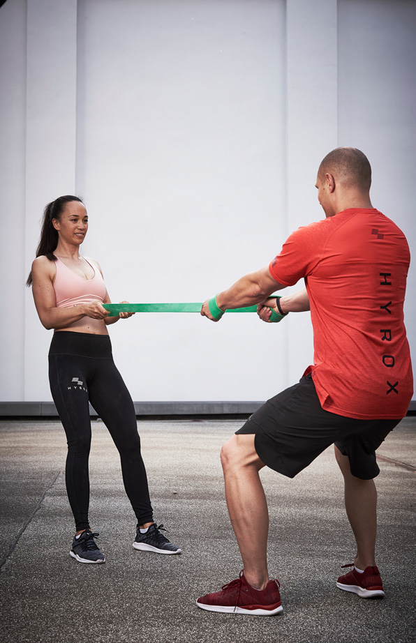 Un homme et une femme effectuant des squat tirés avec une bande pour simuler l'effort du rameur pendant leur entraînement Hyrox.