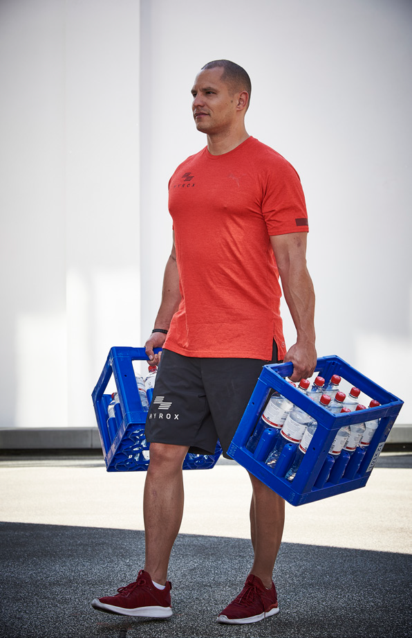 Un homme effectuant des farmer's carry avec des caisses remplies de bouteille pour remplacer les kettlebells pendant son entraînement Hyrox. 